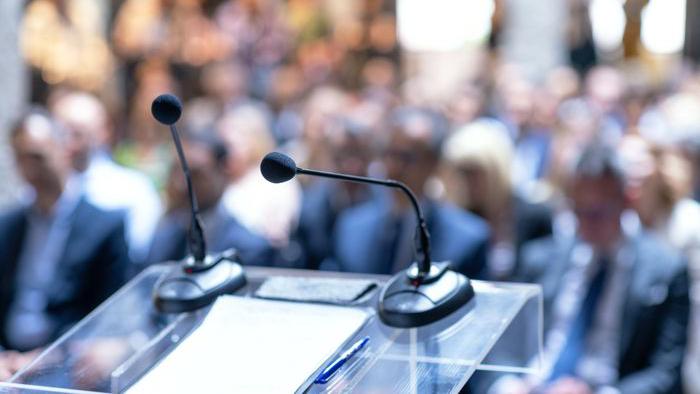 Mikrofoner på ett podium. I fonden syns åhörare